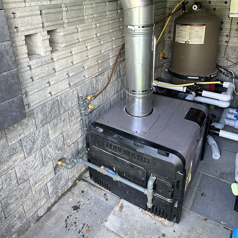 A high-angle shot of a grey and black Hayward pool heater installed inside a stone block utility enclosure. A thick, silver metallic exhaust flue pipe extends vertically from the top of the heater unit. To the left, grey gas lines with yellow shut-off valves are mounted against the textured stone wall. In the background, a large tan cylindrical Hayward pool filter tank connects to an intricate system of white PVC plumbing pipes and valves running along the ground, highlighting a complete residential pool mechanical system.